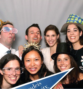 Seven people wearing funny party hats and glasses smile for the camera with a blue Brandeis pennant
